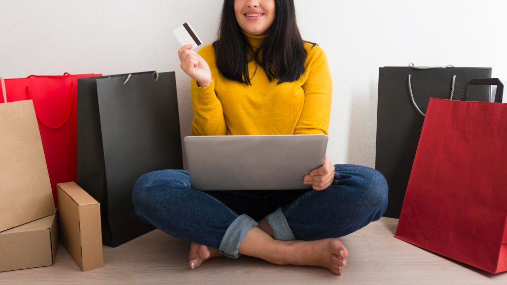 A imagem representa uma mulher com cartão de frente para um computador, com várias sacolas em volta.